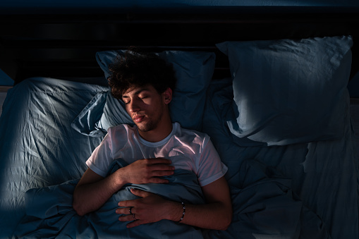 Top view of young man sleeping on a bed in his bedroom at night. Light shining through the window. Healthy sleep, textile bedding. People, bedtime and rest concept. Copy space