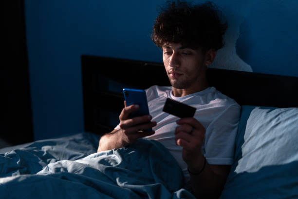 homem bonito sentado na cama e usando o telefone inteligente para pagar com cartão de crédito à noite em casa. cartão bancário de crédito para loja online, entrega de pedidos, reserva de passeio, relaxar em casa no quarto - hotel domestic room telephone technology - fotografias e filmes do acervo