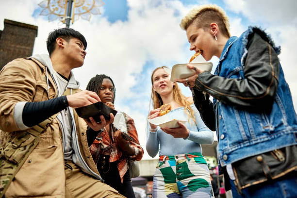 Low angle view of young Londoners enjoying street food Casually dressed Asian, Black, and Caucasian friends in late teens and early 20s eating sandwiches outdoors in East End. london girl stock pictures, royalty-free photos & images