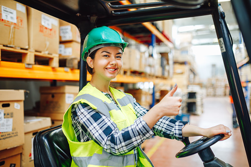 Experience the dynamic atmosphere of a distribution warehouse as a young, happy female forklift driver takes charge of operations. This captivating image showcases her confidence and skill as she poses and smiles while driving the worklift. Embrace the industrial and industrial concept, where efficiency and productivity are paramount. Witness the seamless coordination between the driver and the forklift, reflecting the dedication and expertise necessary in the warehouse environment. This image captures the spirit of success and highlights the pivotal role of skilled workers in ensuring smooth operations.