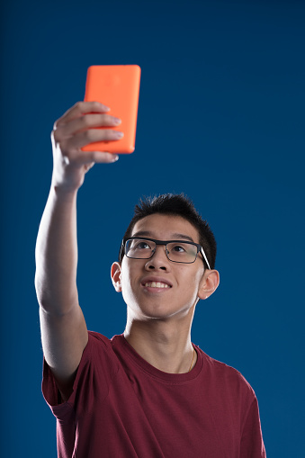 An Asian person takes a selfie with an orange smartphone, or maybe creating content for a live story. He wears glasses and a burgundy shirt