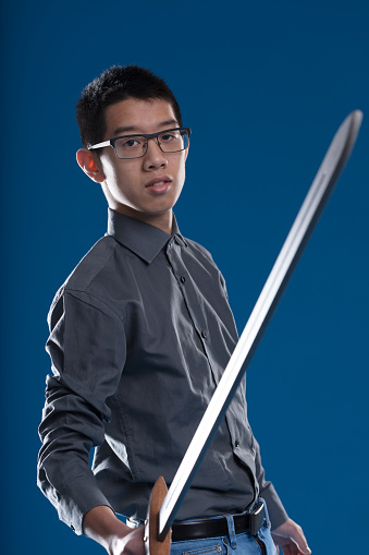 Asian man in workplace attire holds a sword, symbolizing data security and antitheft. He's wearing glasses, with a blue background. Metaphorical and allegorica