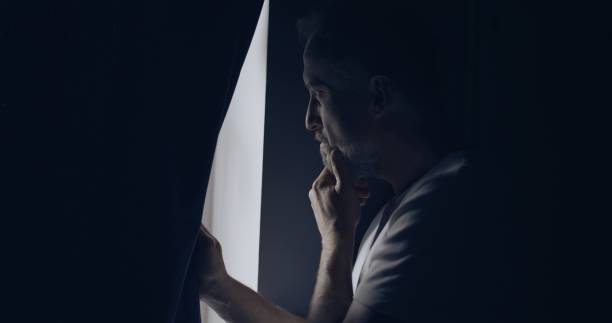 Anxious man alone in a dark room. Looking out the window with worry and touching face stock photo
