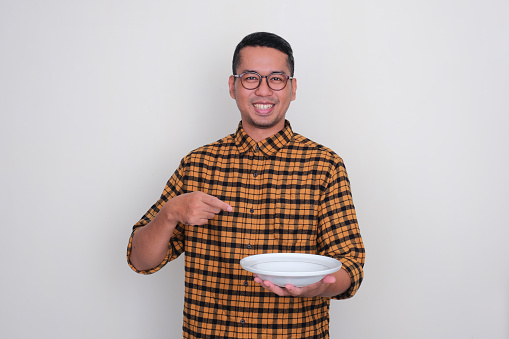 Adult Asian man smiling and ponting to empty dinner plate that he hold