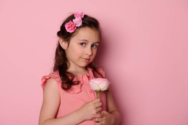 Authentic elegant little girl in pink dress, holding a rose flower, smiles cutely looking at camera, isolated background Beauty portrait of a Caucasian noble charming baby girl in pink dress and flowers in hairstyle, holding pink rose flower, smiling looking at camera, isolated on pink background. Copy ad space cutely stock pictures, royalty-free photos & images