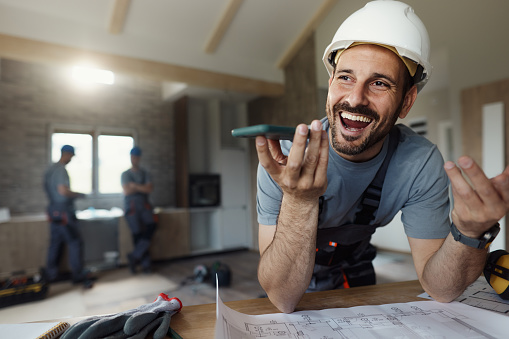 Happy construction worker communicating with someone over mobile phone's speaker while working inside of a house. There are people in the background.