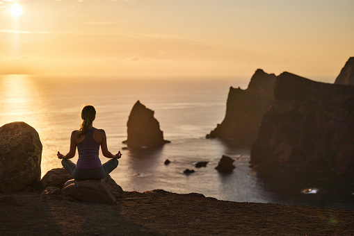 Rear view of athletic woman doing Yoga relaxation exercises on coastline of Ponta De Sao Lourenco at sunset. Copy space.