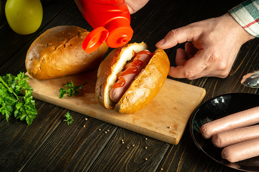 The cook makes a hot dog on the kitchen table with sausages and buns. The chef adds ketchup to the hot dog. Fast food or street food concept