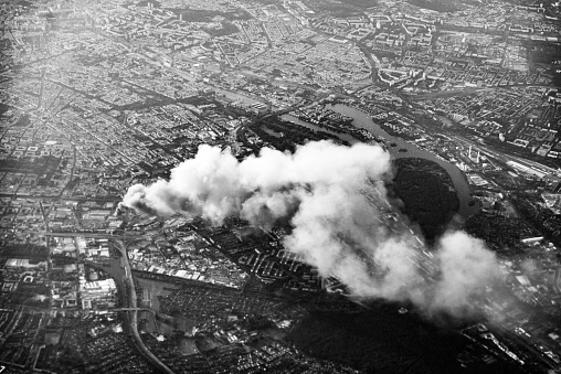 A black and white photo of Berlin from an airplane with a  recycling yard on fire and a column of smoke rising over the city.