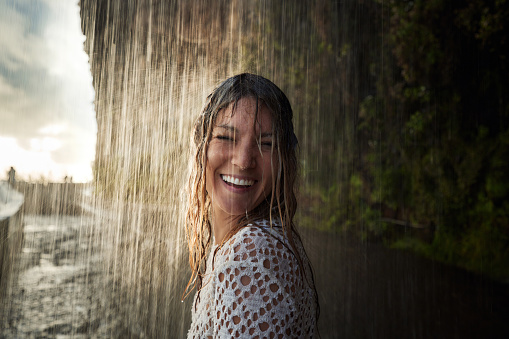 Young wet woman feeling happy while standing under the waterfall in nature.