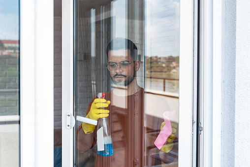 Household and people concept - man in rubber gloves cleaning window with rag and spray cleaner at home. Owner of rental apartment cleaning home for new tenants before they move in as rent agreement.