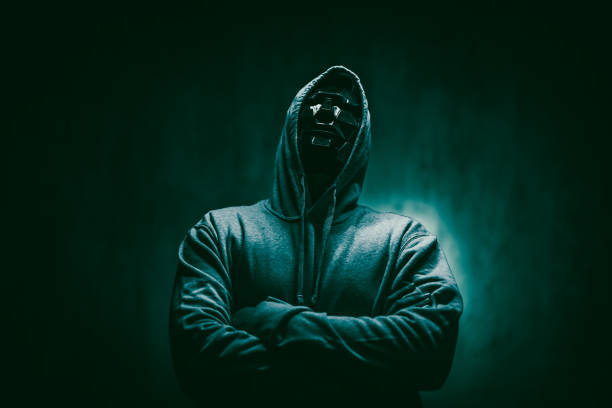Mysterious individual in black hoodie and face mask stares intensely against clean white backdrop.