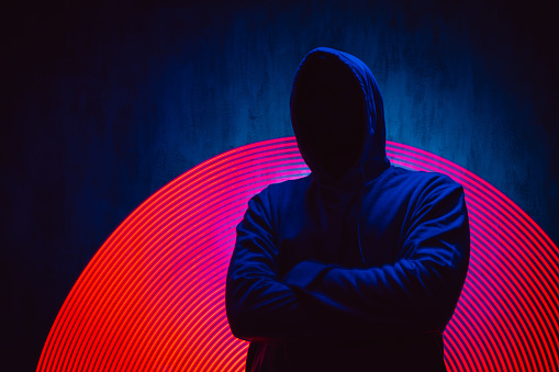 Scary faceless man in a hoodie standing under neon lights, with crossed arms.