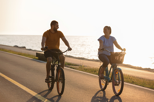 Caucasian couple enjoying a ride on beach cruiser bikes relaxed pedaling and talking near the beautiful sea illuminated by the afternoon sun