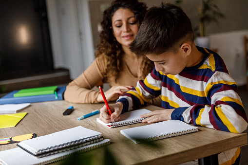 Portrait of diligent boy sitting at table, studying and writing homework with his mother sitting by his side, giving him guidance and assistance.