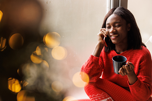 Selective focus shot of happy young Black woman enjoying her morning coffee on the window sill and talking on the phone with a friend, wishing them happy Christmas holidays.