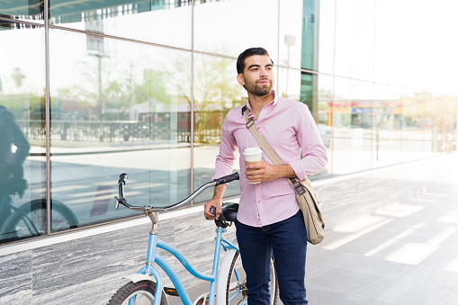 Handsome latin man drinking coffee and smiling while on his commute to work using his bicycle