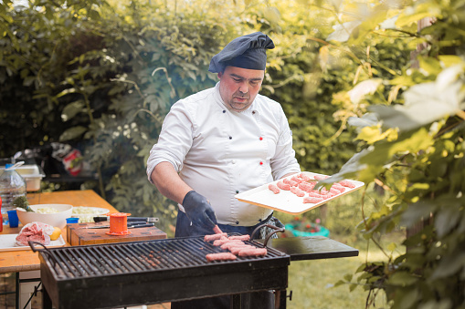 Man wearing apron and chef's hat concentrating onto meat on a grill