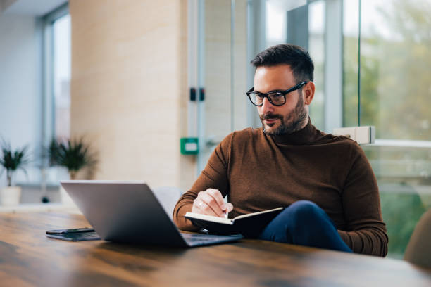 istock Focused businessman looking at the laptop, taking notes in notebook, sitting at the office. 1498934228