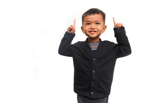 A smiling toddler boy looks at the camera raising his hands and pointing up, isolated on white background