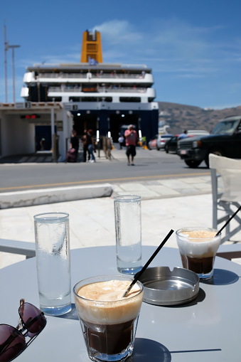 Ios, Greece - May 18, 2021 : Drinking iced Greek coffee, also known as Freddy cappuccino in front of a ferry boat that just arrived at Ios Greece