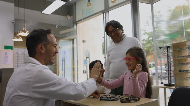 Cheerful optician helping a little girl try on her new glasses and giving her a high five while granddad and brother stand at background