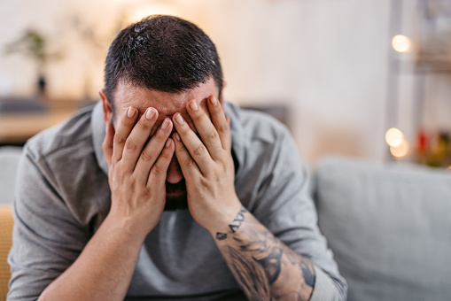 Stressed man sitting alone at home and holding his head covering his face with both hands.