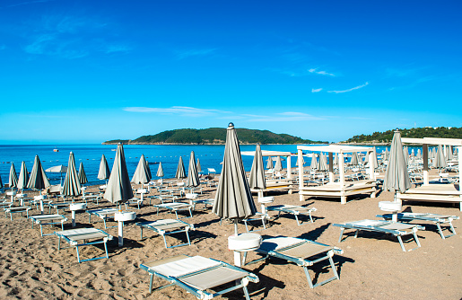 Sun loungers and umbrellas on public beach at summer sunny mornin. Skojl island background. Calm sea and blue sky. Budva Riviera. Seaside vacation resort season. Becici, Montenegro.