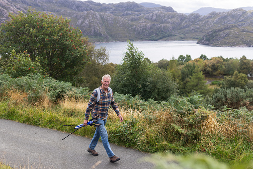 A shot of a senior man walking along a country road beside loch torridon. He is carrying hiking poles and an umbrella in his hands. There is lush foliage and rocky hills in the backdrop.