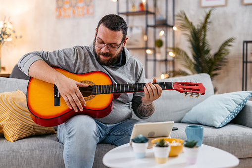 Young overweight man playing an acoustic guitar on the sofa. Watching online tutorials.