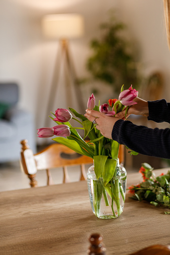 Close up shot of unrecognizable young woman standing over her dining table and arranging tulips and roses in a glass vase to decorate her home with.