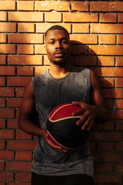 An African American basketball players is standing in front of a brick wall in a gymnasium, holding the ball and looking at the camera.
