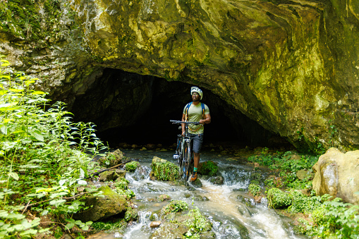 The cyclist comes out of the cave and pushes the bike trough the stream.