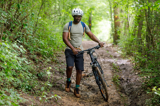 A black cyclist pushes a bicycle over inaccessible terrain in the woods.
