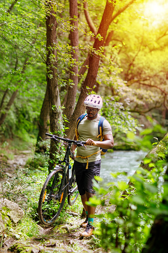 A black cyclist carries a bicycle over inaccessible terrain near to mountain stream.