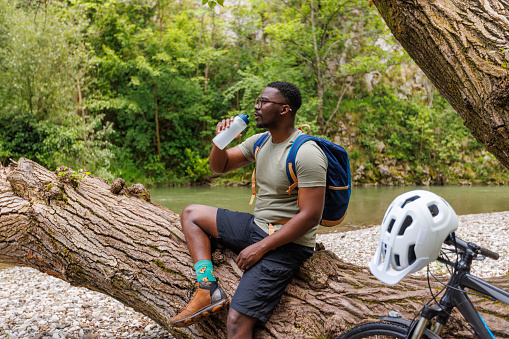 A young black cyclist stands next to his bicycle and looks at a mountain stream.