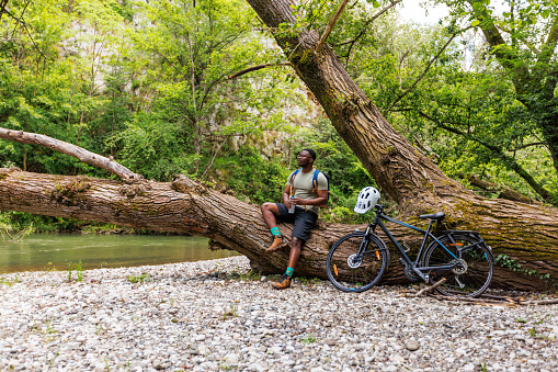 A young black cyclist stands next to his bicycle and looks at a mountain stream.