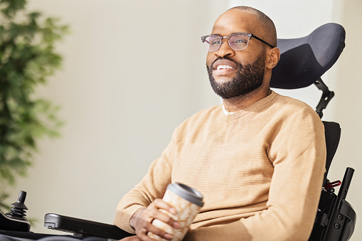 Side view of bearded black male in casual clothes and eyeglasses sitting on wheelchair in living room with a reusable cup