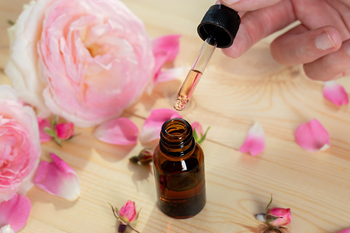 Rose oil in pipette. Against background of roses of pink delicate pastel color and bottle of amber glass