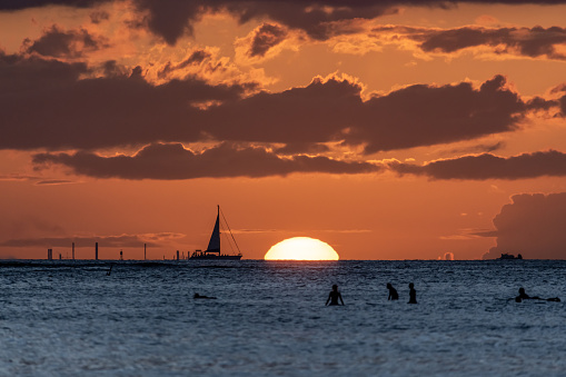 Silhouettes of sail boats and surfers in the pacific ocean off the west coast of Hawaii.