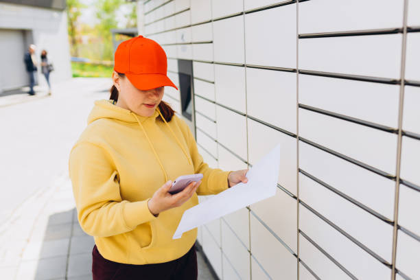 istock Young woman in red hat picks up parcel from automatic post office machine, Courier standing with phone and small box. Concept of fast delivery to automatic self lockers 1498984973