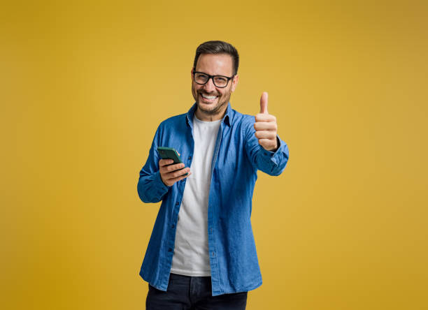 웃고 있는 기업가의 초상화는 엄지손가락을 치켜세우고 노란색 배경에 스마트폰으로 메시지를 보낸다 - men cheerful happiness smart phone 뉴스 사진 이미지