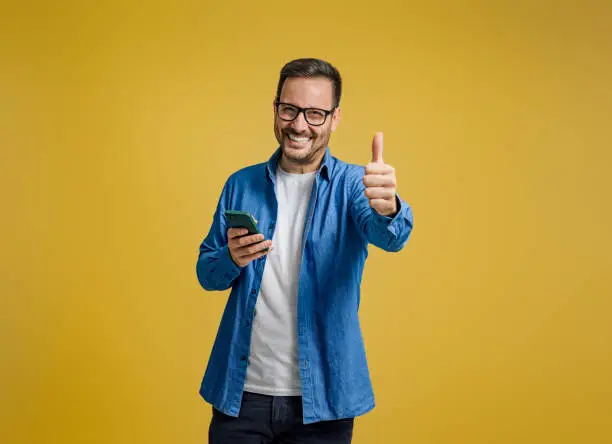 Photo of Portrait of smiling entrepreneur showing thumbs up and messaging on smartphone on yellow background