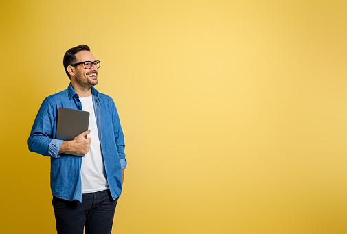 Happy confident male entrepreneur holding digital tablet and contemplating over yellow background