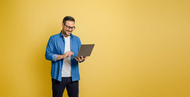 hombre de negocios sonriente con anteojos trabajando en una computadora portátil mientras está de pie sobre fondo amarillo - businessman happiness carefree computer fotografías e imágenes de stock