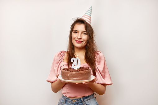 The girl is holding a festive cake with a candle in the form of the number 20. The concept of a birthday celebration.