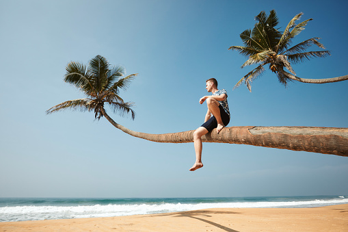 Man sitting on trunk of palm tree and enjoying sea view. Idyliic sand beach in Sri Lanka.