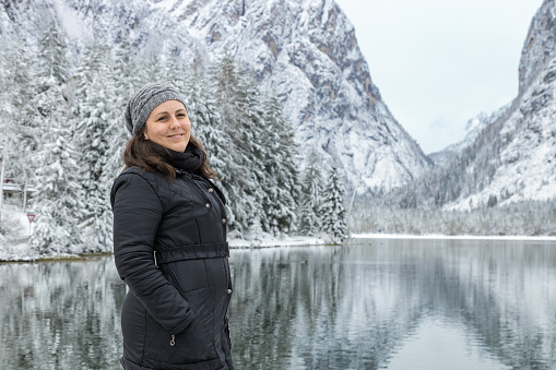 Woman in black clothes posing and smiling by a beautiful view of Toblacher See (Dobbiaco Lake) in a snowy winter day. Reflections of mountains and trees on water; Dolomites, Bolzano, Italy
