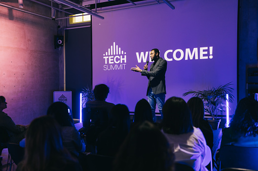In a dynamic setting of a startup tech conference, a business entrepreneur stands before a captivated audience, delivering an inspiring presentation. Their passionate delivery and insightful perspectives resonate with the attendees.