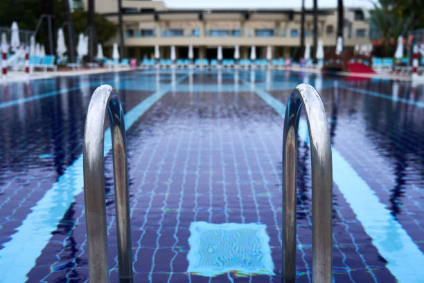 Metal pool ladder with blurred swimming pool and closed umbrellas in background on a cloudy day